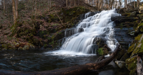 A serene waterfall cascades over rocks into a calm pool, surrounded by trees and lush greenery.
