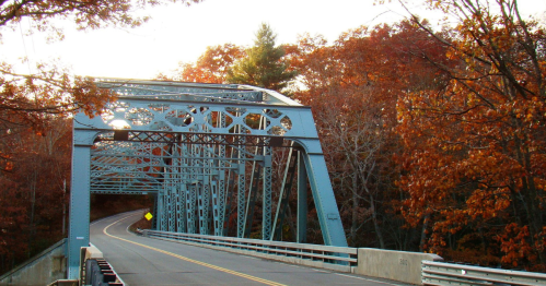 A blue metal bridge spans a winding road, surrounded by autumn foliage in warm orange and yellow hues.