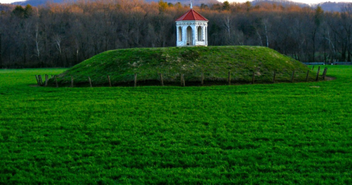 A small white gazebo atop a grassy mound, surrounded by a wooden fence, with trees in the background.