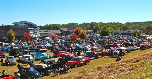 A large crowd at a car show with numerous classic cars displayed on a sunny day, surrounded by trees and buildings.