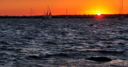 A serene sunset over water, with sailboats silhouetted against the vibrant orange and yellow sky.