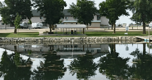A calm lake reflects trees and a pavilion, with a building visible in the background.