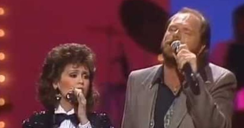 A man and woman sing together on stage, with colorful lights and a band in the background.