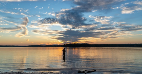A person stands in shallow water at sunset, with vibrant clouds reflecting on the calm lake surface.