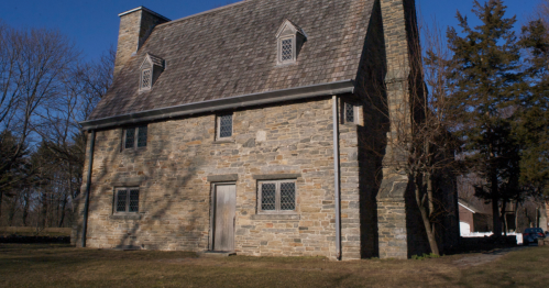 A historic stone building with a sloped roof, featuring multiple windows and surrounded by trees and grass.