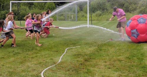 Children spray water at a large inflatable soccer ball while a man directs the water, all on a grassy field.