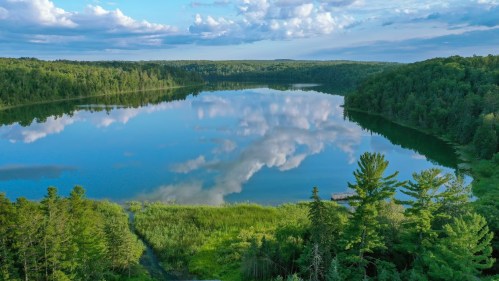 A serene lake surrounded by lush greenery and trees, reflecting clouds in the calm water under a blue sky.