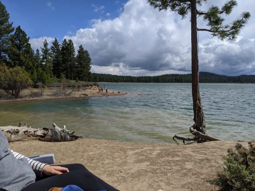 A serene lakeside scene with trees, clouds, and people walking along the shore.