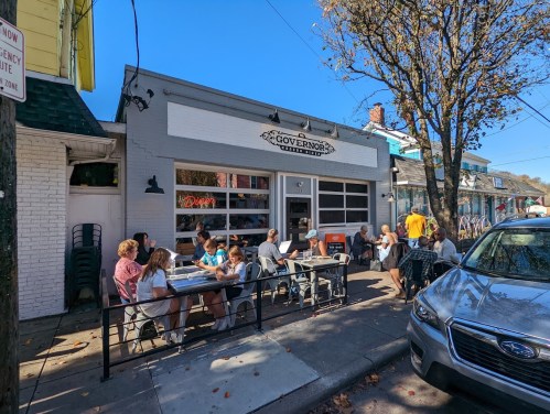 A bustling café with outdoor seating, patrons enjoying meals, and a clear blue sky overhead.