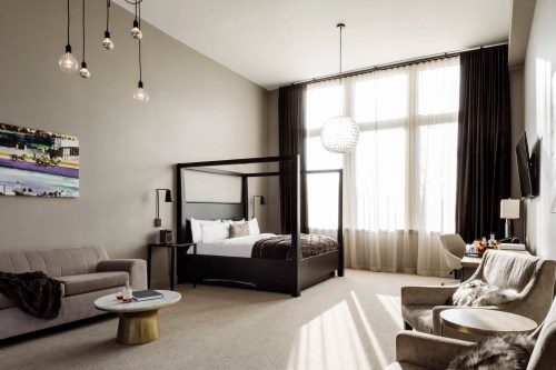 Modern hotel room featuring a four-poster bed, stylish seating, large windows, and contemporary decor.