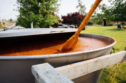 A large metal container filled with orange liquid, with a wooden paddle stirring it, set in a grassy outdoor area.