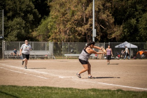 A woman pitches a softball while a runner approaches first base in a sunny park setting. Spectators are in the background.