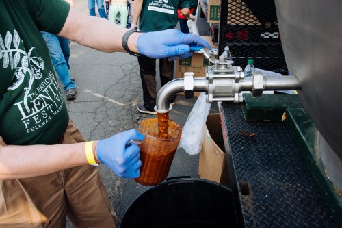A person in gloves pours a brown liquid from a spout into a measuring cup at a festival.