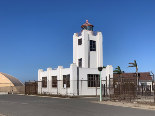 A white, art deco-style building with a tower, surrounded by a fence and palm trees, under a clear blue sky.