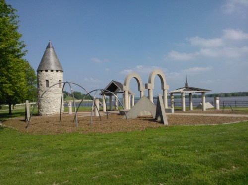 A playground featuring a castle tower, arches, and climbing structures, set against a lake and blue sky.