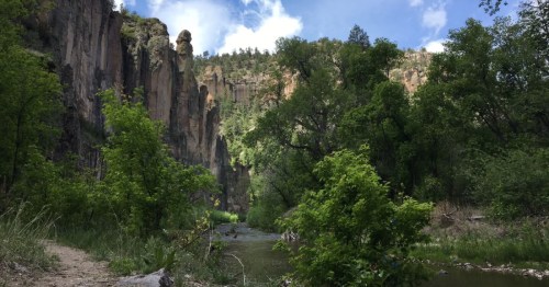 A serene landscape featuring a river surrounded by lush greenery and towering cliffs under a blue sky with clouds.