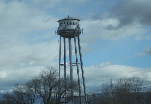 Water tower with "Los Lunas" written on it, surrounded by trees and a cloudy sky.