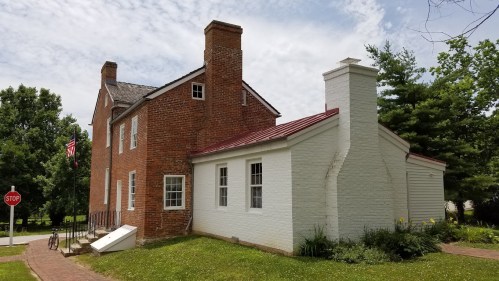 A two-story brick house with a red metal roof, featuring a white side extension and surrounded by greenery.