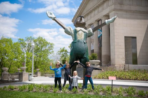 Four children pose playfully in front of a large statue with outstretched arms, set against a bright blue sky.