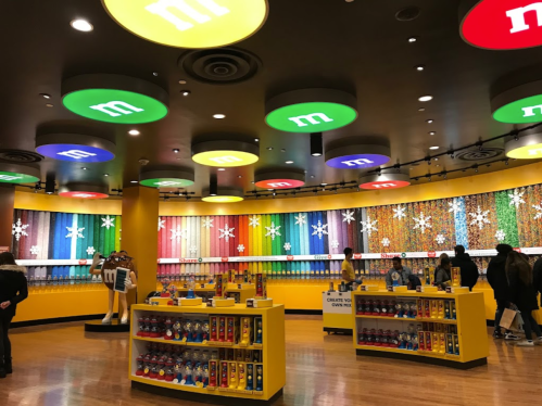Colorful M&M's store interior with vibrant ceiling lights, candy displays, and customers browsing.