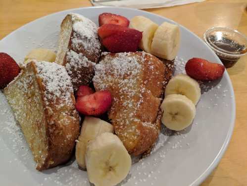 A plate of French toast topped with powdered sugar, strawberries, and banana slices, served with a side of syrup.