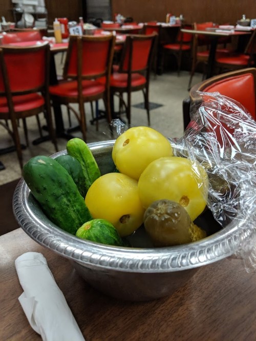 A silver bowl filled with yellow tomatoes, cucumbers, and pickles, placed on a wooden table in a diner.