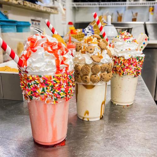 Three colorful milkshakes topped with whipped cream, sprinkles, and various candies, served in clear cups with straws.