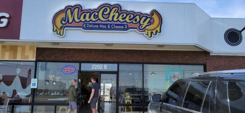 Exterior of MacCheesy restaurant with a colorful sign, featuring two people outside and a parked vehicle.