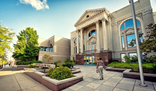A person walks towards a grand building with classical architecture, surrounded by greenery and a clear blue sky.