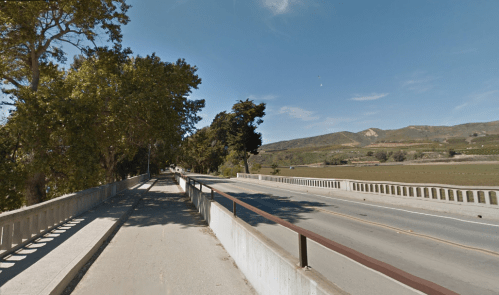 A wide, empty road alongside a concrete barrier, lined with trees and hills in the background under a clear blue sky.