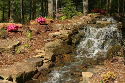 A serene waterfall cascades over rocks, surrounded by lush greenery and vibrant pink flowers in a tranquil garden setting.