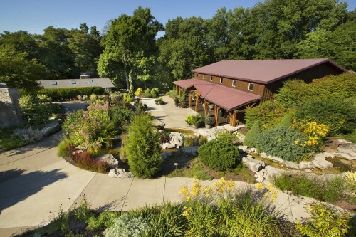 A scenic view of a landscaped garden with a wooden building and pathways surrounded by lush greenery and colorful flowers.