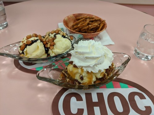 Two ice cream sundaes in glass dishes, topped with nuts, chocolate sauce, caramel, and whipped cream, with a bowl of pretzels.