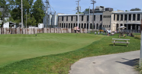 A green golf course with a path, picnic tables, and a building in the background, featuring people enjoying the outdoors.
