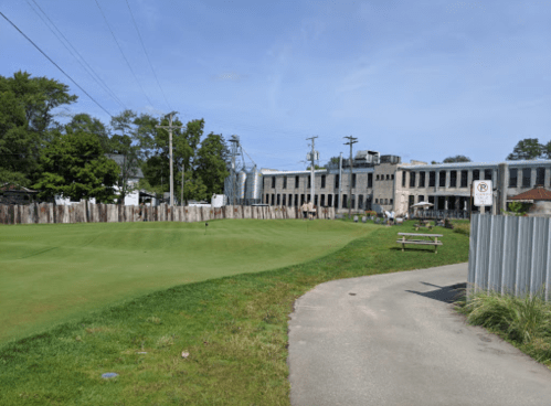 A grassy area with a path, leading to a building and trees, with people in the background enjoying the space.