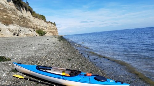A blue kayak rests on a rocky beach with cliffs and calm water under a clear blue sky.