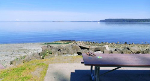 A serene lakeside view with calm water, a picnic table, and distant hills under a clear blue sky.