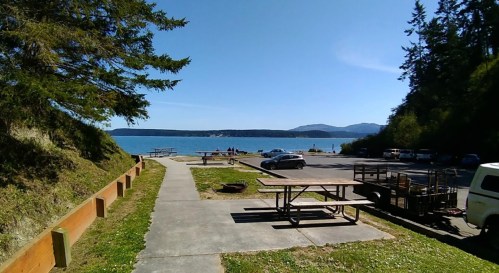 A scenic view of a waterfront area with picnic tables, a parking lot, and distant mountains under a clear blue sky.