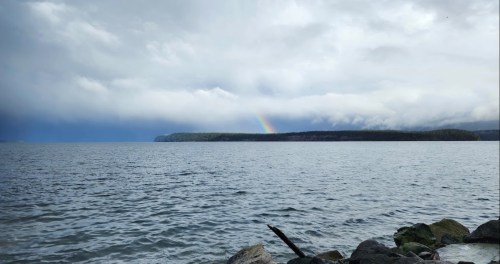A serene seascape with gray clouds and a faint rainbow arching over a distant shoreline. Rocks line the water's edge.