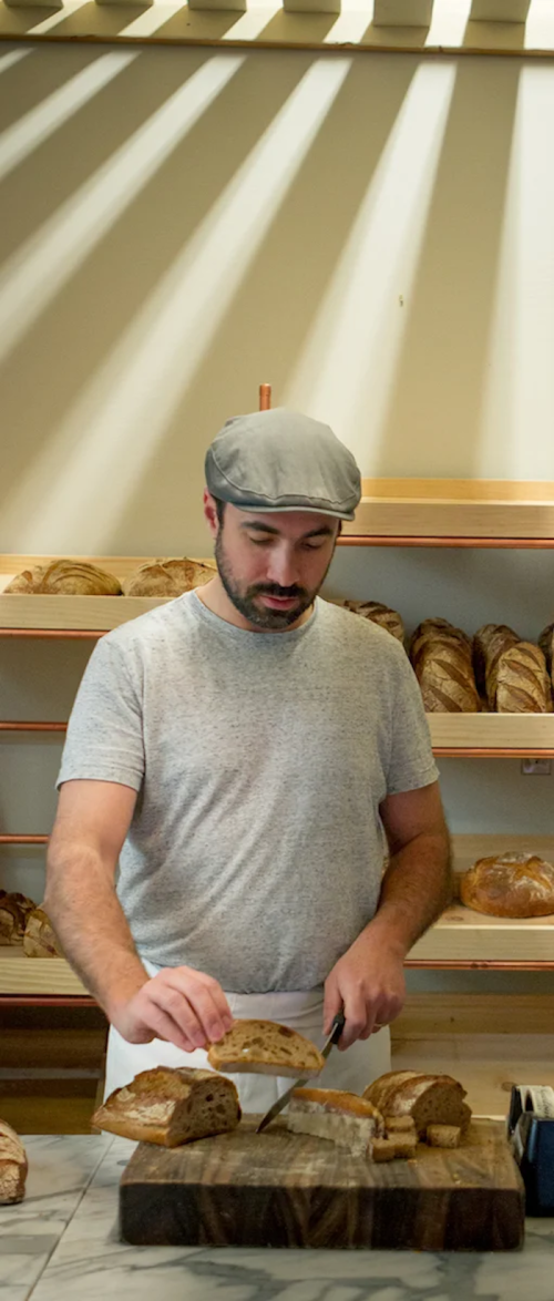 A man in a gray cap slices bread in a bakery with shelves of various loaves in the background.