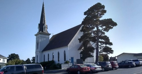 A white church with a tall steeple, surrounded by parked cars and a large tree, under a clear blue sky.