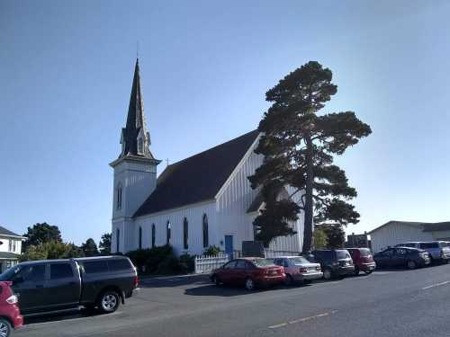 A white church with a tall steeple, surrounded by parked cars and a large tree, under a clear blue sky.