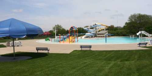 A sunny outdoor pool area featuring water slides, colorful play structures, and shaded seating on green grass.