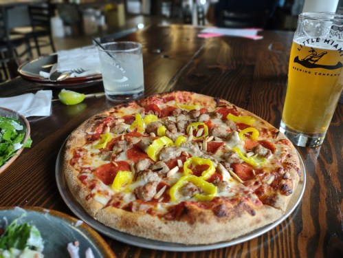 A pizza topped with sausage, peppers, and onions, served with a beer on a wooden table in a casual dining setting.