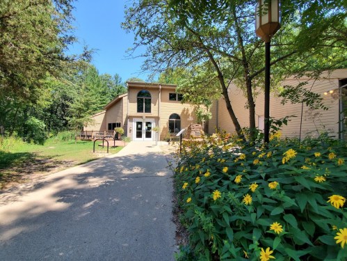 A pathway leads to a light-colored building surrounded by greenery and yellow flowers under a clear blue sky.