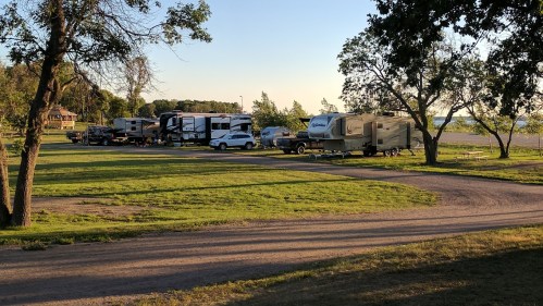 A campsite with several RVs parked on a grassy area near a lake, surrounded by trees and a clear blue sky.