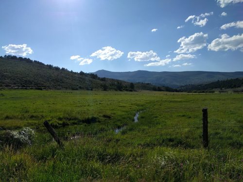 A scenic landscape featuring a green field, distant mountains, and a blue sky with scattered clouds.