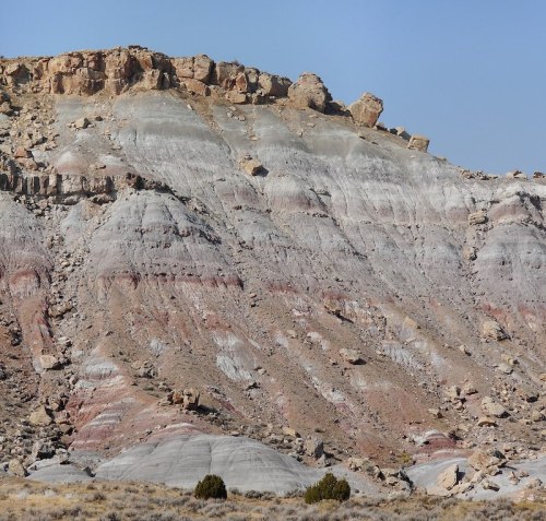 Layered rock formations in shades of gray, red, and beige, with a rocky cliff and sparse vegetation in the foreground.