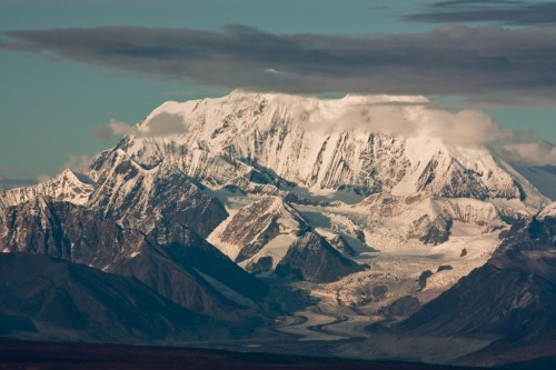 Snow-capped mountain peaks rise above a glacier, surrounded by rugged terrain and a cloudy sky.