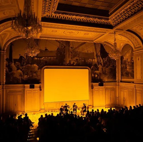 A dimly lit theater with a large screen and a band performing in front of an audience, surrounded by ornate decor.
